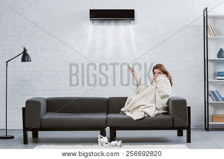 Young Woman Covered With Blanket On Couch Under Air Conditioner Hanging On Wall And Blowing Cooled A