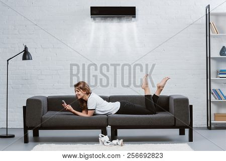 Happy Young Woman Using Smartphone On Couch Under Air Conditioner Hanging On Wall