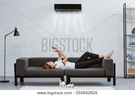 Relaxed Young Woman Using Smartphone On Couch Under Air Conditioner Hanging On Wall