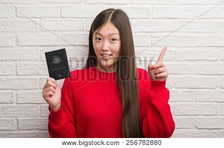 Young Chinese woman over brick wall holding passport of Australia surprised with an idea or question pointing finger with happy face, number one