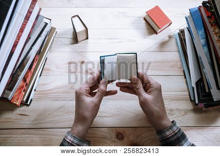 A Man Reading A Miniature Book Among The Big Books In The Library At The Wooden Table.