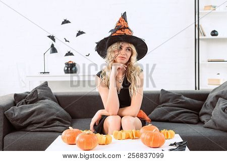 Portrait Of Pensive Woman In Witch Costume Sitting At Coffee Table With Pumpkins, Halloween Holiday 