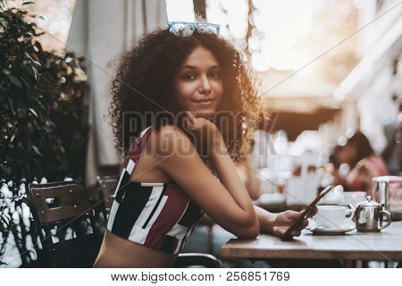 Cute Curly African-american Female In A Crowded Street Bar Holding A Smartphone, With The Cup Of A H
