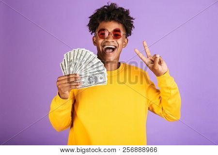 Portrait of stylish african american man wearing sunglasses smiling while holding dollars money banknotes isolated over violet background