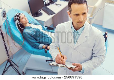 Serious Doctor Taking Notes During Electroencephalography Procedure