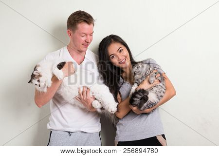 Young Happy Multi-ethnic Couple Holding Two Cats Together Against White Background