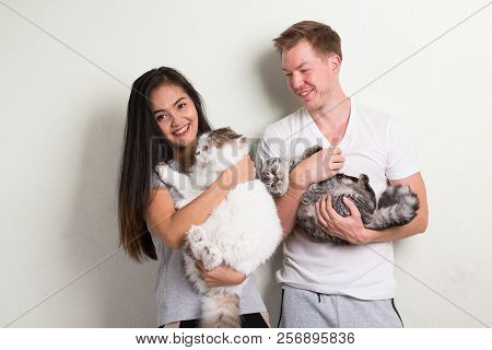 Young Happy Multi-ethnic Couple Holding Two Cats Together Against White Background