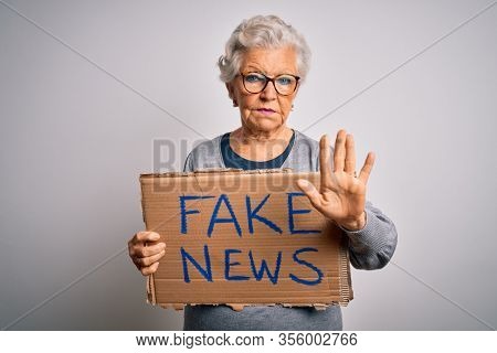 Senior grey-haired woman holding banner with fake news message over white background with open hand doing stop sign with serious and confident expression, defense gesture