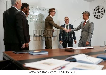 Caucasian And Asian Businessmen In Classic Wear Shaking Hands After Signing The Agreement While Stan