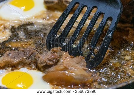 Fried Eggs, Black Spatula Turns Over Pieces Of Meat In A Pan In Sunflower Oil Close-up. The Concept 