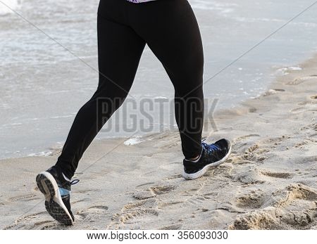Legs Of A Runner Running At The Beach In Black Spandex At The Edge Of The Water On The Sand.