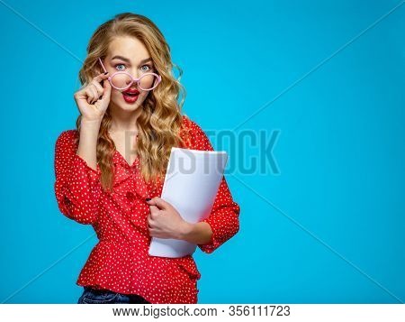 Surprised businesswoman in a casual clothes holds papers, at studio. Shocked woman secretary in a red shirt over blue background holding documents.  Female student in a pink glasses holding documents 