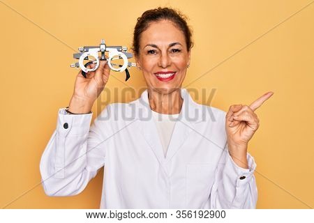 Middle age senior oculist woman holding optometrical glasses for vision correction very happy pointing with hand and finger to the side