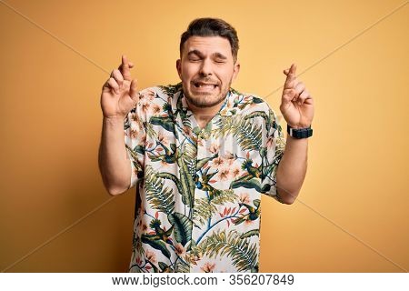 Young man with blue eyes on vacation wearing floral summer shirt over yellow background gesturing finger crossed smiling with hope and eyes closed. Luck and superstitious concept.