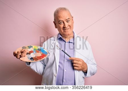 Middle age artist man painting using paintbrush and palette with colors over pink background with a happy face standing and smiling with a confident smile showing teeth