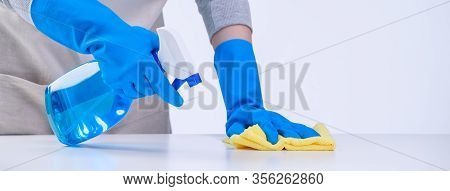 Young Woman Housekeeper In Apron Is Cleaning, Wiping Down Table Surface With Blue Gloves, Wet Yellow