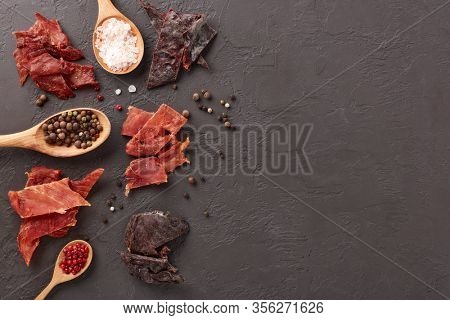 Jerky. Set of various kind of dried spiced meat, diverse peppercorns and salt on wooden spoons  on dark gray background. Top view with copy space. Snack for beer.