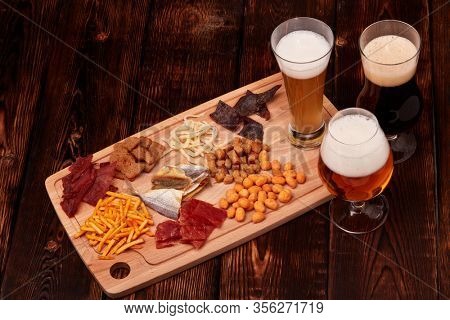Three glasses with different beer and set of various kind snacks: jerky,  potato chips, salted nuts, stockfish, bread spicy crackers, dried squids on wooden table on brown background. Snacks for beer.