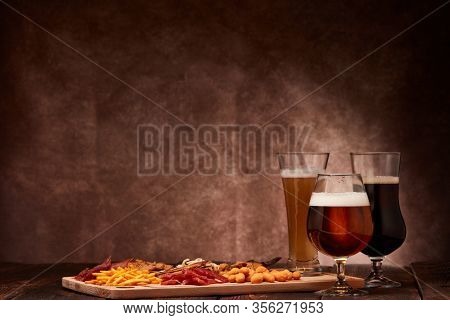 Three glasses with different beer and set of various kind snacks: jerky,  potato chips, salted nuts, stockfish, bread spicy crackers, dried squids on wooden table on brown background. Snacks for beer.