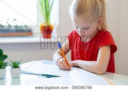 Serios Blonde Schoolgirl Sitting At Home And Doing School Homework, Writing In Notebook. Training Bo