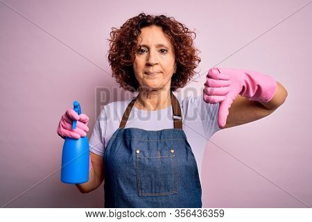 Middle age curly hair woman cleaning doing housework wearing apron and gloves using spayer with angry face, negative sign showing dislike with thumbs down, rejection concept