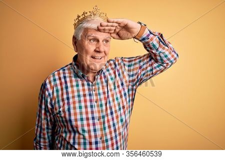 Senior handsome hoary man wearing golden crown of king over isolated yellow background very happy and smiling looking far away with hand over head. Searching concept.