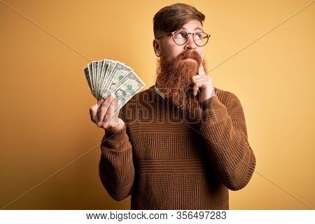 Redhead Irish man with beard holding a bunch of 1 dolla banknotes over yellow background serious face thinking about question, very confused idea