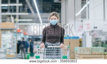 A Woman In A Medical Mask Stands In A Supermarket With A Timelapse Grocery Cart. Protection From Cor