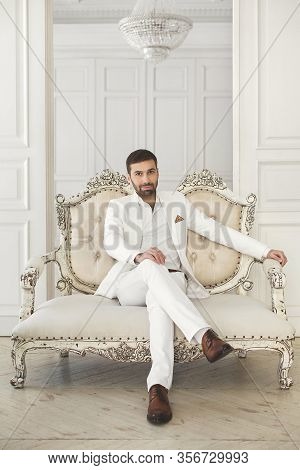 Elegant Young Handsome Man With A Beard In A White Classic Suit. The Hotels Interior. Photo Studio. 