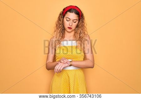 Beautiful blonde pin-up woman with blue eyes wearing diadem standing over yellow background Checking the time on wrist watch, relaxed and confident