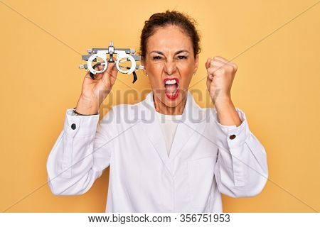 Middle age senior oculist woman holding optometrical glasses for vision correction annoyed and frustrated shouting with anger, crazy and yelling with raised hand, anger concept