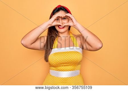 Young hispanic pin up woman wearing fashion sexy 50s style over yellow background Doing heart shape with hand and fingers smiling looking through sign