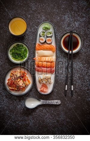 Asian food assortment. Various sushi rolls placed on ceramic plates. Kimchi and goma wakame salads. Soy souce and chopsticks on sides. Grungy dark background with copy space.