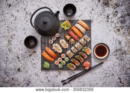 Assortment of different kinds of sushi rolls placed on black stone board. Traditional asian iron tea pot on side. Top angle view.