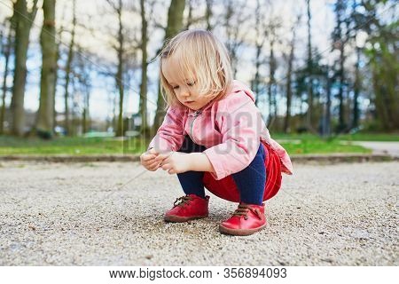 Adorable Baby Girl In Park, Sitting On Hunkers And Playing With Little Stones. Little Child Having F