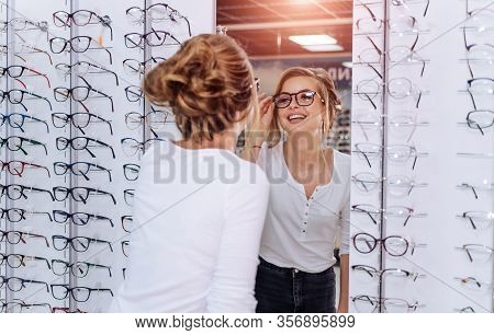 Happy Female Optometrist, Optician Is Standing With Many Glasses In Background In Optical Shop. Stan