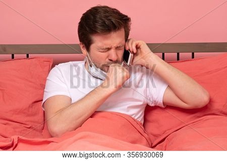 Sick Young Cauacsian Man Coughing While Lying In Bed And Speaking On Phone With Doctor.