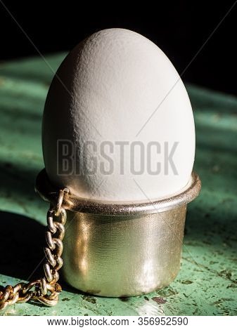 A White Egg Stands In A Metal Egg Stand On The Table, On A Black Background, Close- Up. Silver Egg S