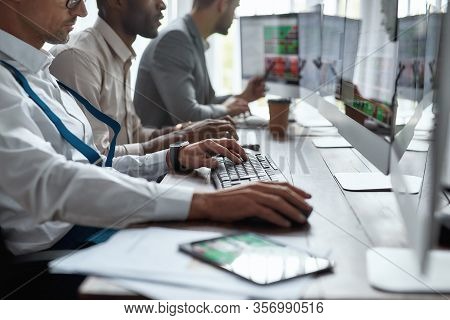 Three Businessmen Sitting At Desktop Front Pc Computers With Financial Graph And Statistics On Monit