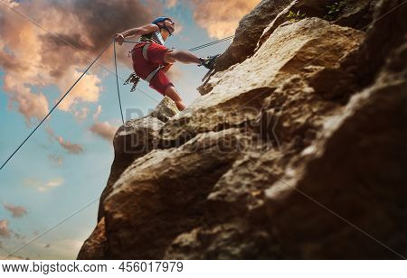 Muscular Climber Man In Protective Helmet Abseiling From Cliff Rock Wall Using Rope Belay Device And