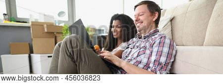 Low Angle Of Cheerful Smiling Couple Watch Show Online On Laptop At Home. Man And Latin Girl Sit On 