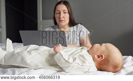 Mother Freelancer Looks After Baby Girl While Working On Laptop In Bedroom. Newborn Child Wiggles Le