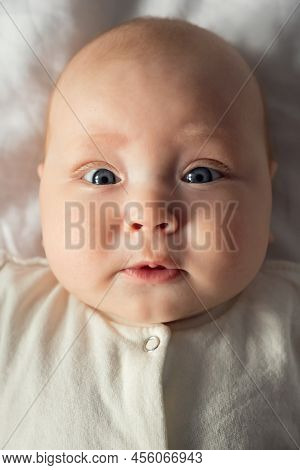 Cute Face Of Little Baby Girl With Large Light Grey Eyes. Newborn Lies On White Sheets And Wiggles A