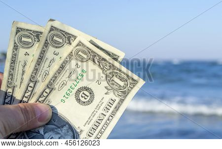 Person Holds In His Hand Three Dollar Bills On The Background Of The Waves Of The Blue Sea. Concept 