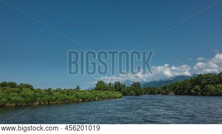 The Bed Of A Calm River Bends. A Tiny Rafting Boat With People Is Visible.lush Green Vegetation On T