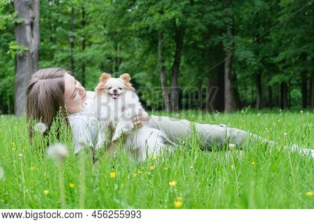 Young Woman With Long Hair Lies On The Lawn In The Park With A Pomeranian Dog