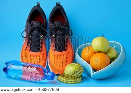 Orange Sneakers, Measuring Tape, Water Botlle And Fruits Apples And Oranges On A Blue Background. Fi
