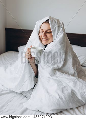 Young Happy Redhead Woman With Cup Of Coffee Sitting In Bed Under Blanket At Home Bedroom. Comfort O