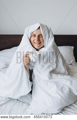 Young Happy Redhead Woman With Cup Of Coffee Sitting In Bed Under Blanket At Home Bedroom. Comfort O