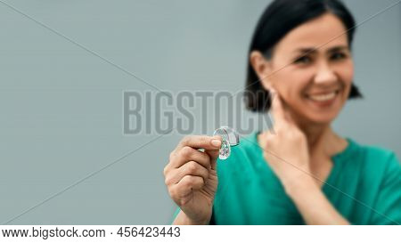 Mature Woman Holding Bte Hearing Aid In Hand On Foreground And Points To Her Ear. Treatment Of Deafn